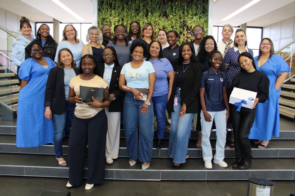 Diverse group of professionals posing on stairs in a bright lobby, some holding folders or boxes and smiling at the camera.