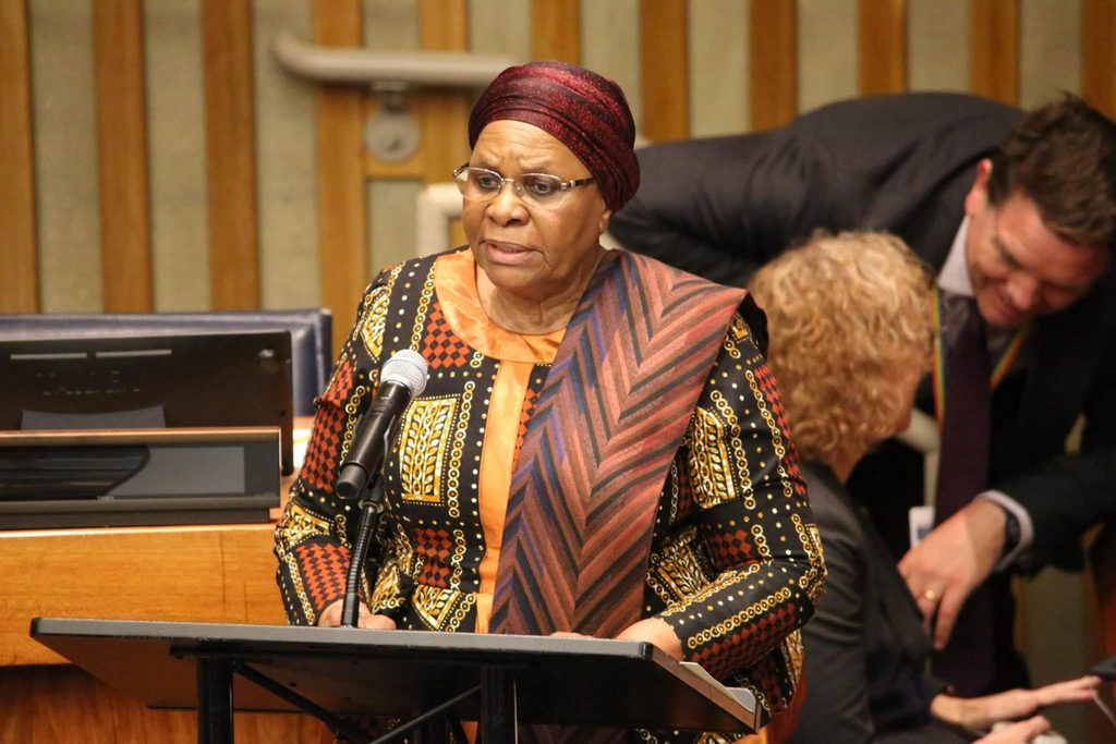 Woman in colorful traditional attire speaks at a podium with a microphone in a conference room.
