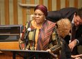 Woman in colorful traditional attire speaks at a podium with a microphone in a conference room.