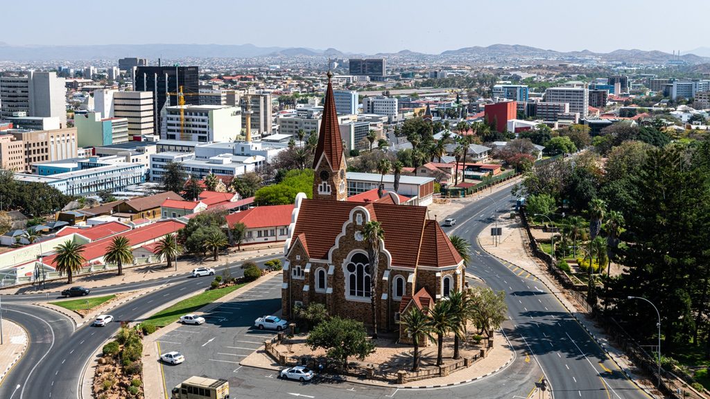 WINDHOEK3 - The Brief | Namibia's Leading Business & Financial News Aerial view of a city with a red-brick church at a roundabout center, surrounded by roads and palm trees.