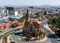 WINDHOEK3 - The Brief | Namibia's Leading Business & Financial News Aerial view of a city with a red-brick church at a roundabout center, surrounded by roads and palm trees.