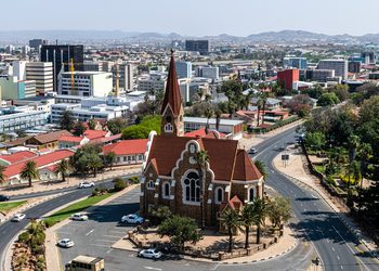 WINDHOEK3 - The Brief | Namibia's Leading Business & Financial News Aerial view of a city with a red-brick church at a roundabout center, surrounded by roads and palm trees.