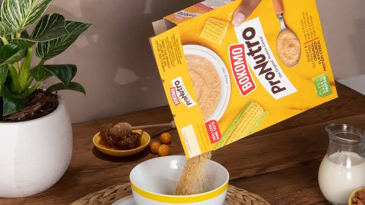 Cereal box tilted as its contents pour into a white bowl with a yellow rim on a wooden table.