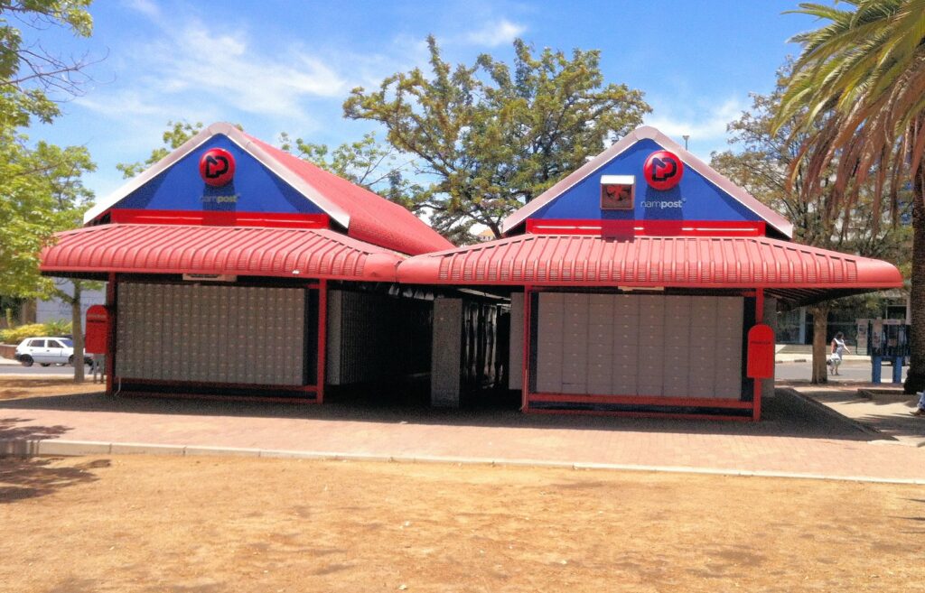 NAMPOST1 - The Brief | Namibia's Leading Business & Financial News Two red-roofed NamPost post office kiosks with shuttered fronts in a sunny outdoor plaza, blue gable signs above.
