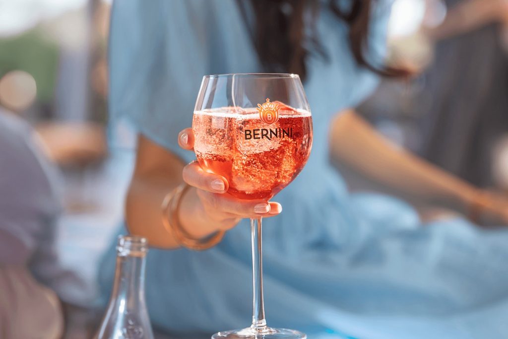 BERNINI - The Brief | Namibia's Leading Business & Financial News Person holding a stemmed glass of pink sparkling drink with the Bernini logo outdoors.