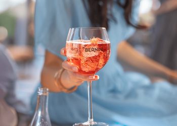 Person holding a stemmed glass of pink sparkling drink with the Bernini logo outdoors.