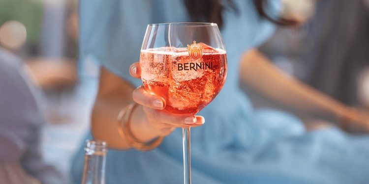 Person holding a stemmed glass of pink sparkling drink with the Bernini logo outdoors.