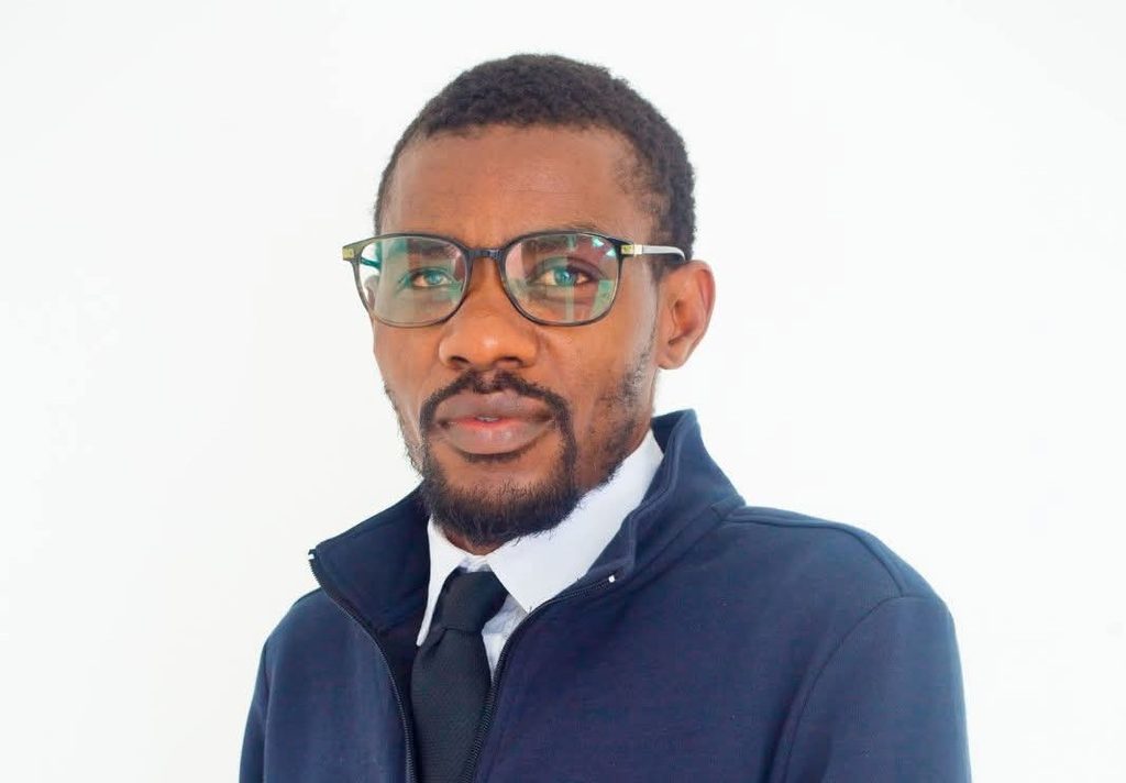 Professional headshot: man with glasses, white shirt, black tie, and navy jacket facing the camera against a white background.