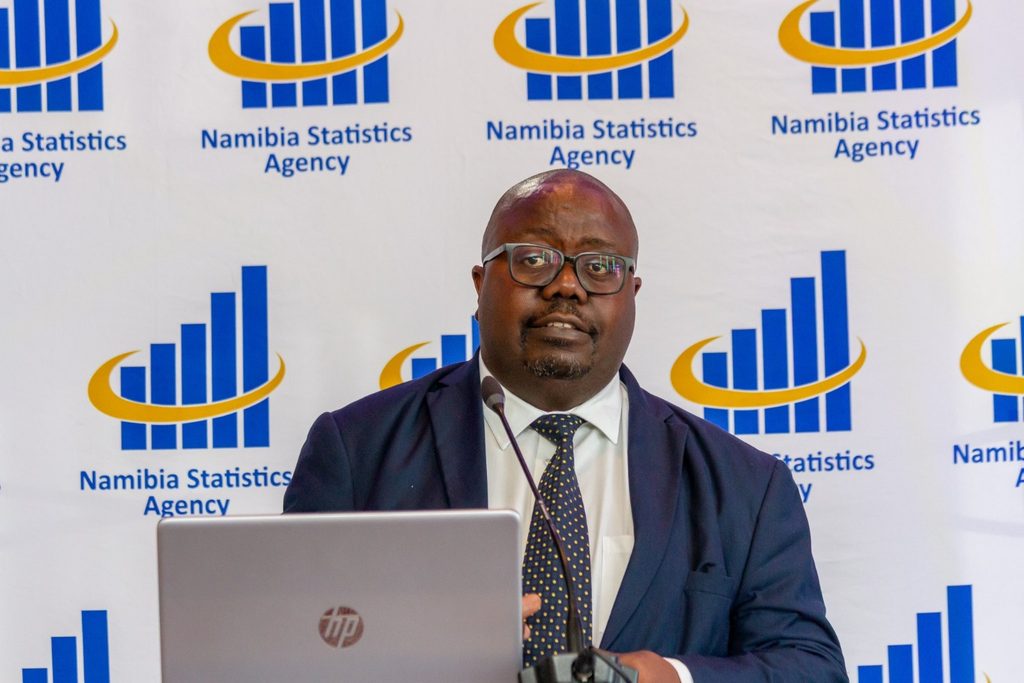 Man in a navy suit and polka-dot tie speaking at a podium in front of a Namibia Statistics Agency backdrop with a laptop in the foreground.