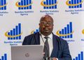 Man in a navy suit and polka-dot tie speaking at a podium in front of a Namibia Statistics Agency backdrop with a laptop in the foreground.