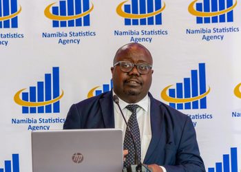 Man in a navy suit and polka-dot tie speaking at a podium in front of a Namibia Statistics Agency backdrop with a laptop in the foreground.