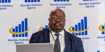 Man in a navy suit and polka-dot tie speaking at a podium in front of a Namibia Statistics Agency backdrop with a laptop in the foreground.