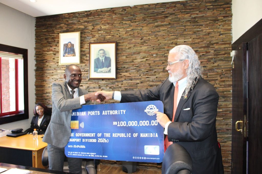 WhatsApp Image 2026-09-30 at 94805 AM - The Brief | Namibia's Leading Business & Financial News Two men in suits shake hands while presenting a blue ceremonial cheque from the Namibian Ports Authority in an office with a stone wall and framed portraits.