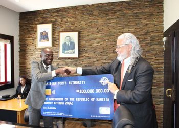Two men in suits shake hands while presenting a blue ceremonial cheque from the Namibian Ports Authority in an office with a stone wall and framed portraits.