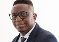 MOSES - The Brief | Namibia's Leading Business & Financial News Professional head-and-shoulders portrait of a man wearing a navy suit, light blue shirt, and tie, smiling at the camera with glasses.