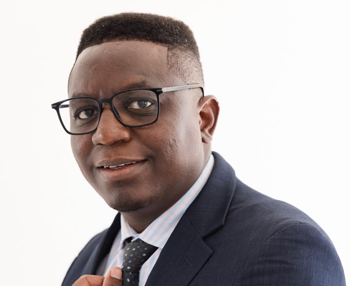 MOSES - The Brief | Namibia's Leading Business & Financial News Professional head-and-shoulders portrait of a man wearing a navy suit, light blue shirt, and tie, smiling at the camera with glasses.