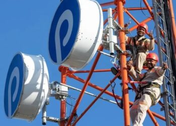 Two technicians in hard hats climb a red telecom tower with large white circular antennas against a clear blue sky.