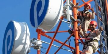 Two technicians in hard hats climb a red telecom tower with large white circular antennas against a clear blue sky.