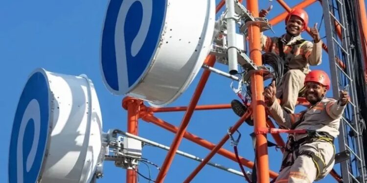 Two technicians in hard hats climb a red telecom tower with large white circular antennas against a clear blue sky.