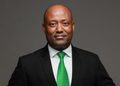Portrait of a man in a black suit and green tie, facing the camera against a gray backdrop.