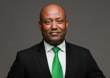 Portrait of a man in a black suit and green tie, facing the camera against a gray backdrop.