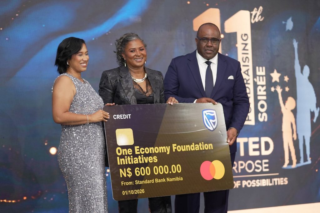 Three formally dressed people pose on stage holding a large novelty cheque reading 'One Economy Foundation Initiatives' for N0,000 from Standard Bank Namibia, dated 01/10/2026, with a blue-lit background.