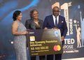 Three formally dressed people pose on stage holding a large novelty cheque reading 'One Economy Foundation Initiatives' for N$600,000 from Standard Bank Namibia, dated 01/10/2026, with a blue-lit background.