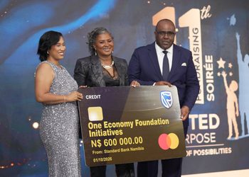 Three formally dressed people pose on stage holding a large novelty cheque reading 'One Economy Foundation Initiatives' for N$600,000 from Standard Bank Namibia, dated 01/10/2026, with a blue-lit background.