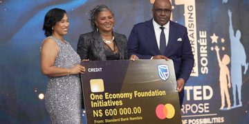 Three formally dressed people pose on stage holding a large novelty cheque reading 'One Economy Foundation Initiatives' for N$600,000 from Standard Bank Namibia, dated 01/10/2026, with a blue-lit background.