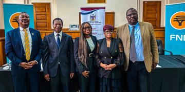 Five professionals in formal attire posing for a photo in a conference room, with banners in the background on either side.