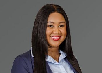 Portrait of a smiling professional woman with long dark hair, wearing a navy blazer and light blue blouse against a gray background (informative)