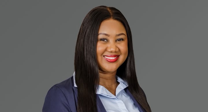 Portrait of a smiling professional woman with long dark hair, wearing a navy blazer and light blue blouse against a gray background (informative)