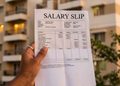 A hand holding a salary slip labeled 'SALARY SLIP' outdoors with apartment buildings in the background