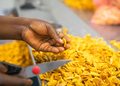 Close-up of a hand picking a yellow snack chip from a pile of fried chips on a production belt.
