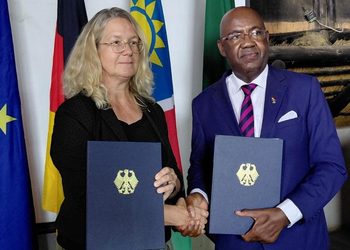 WhatsApp Image 2026-10-06 at 113001 AM - The Brief | Namibia's Leading Business & Financial News Two professionals shake hands while each holds a navy folder with a gold emblem, standing before national flags in a formal setting.