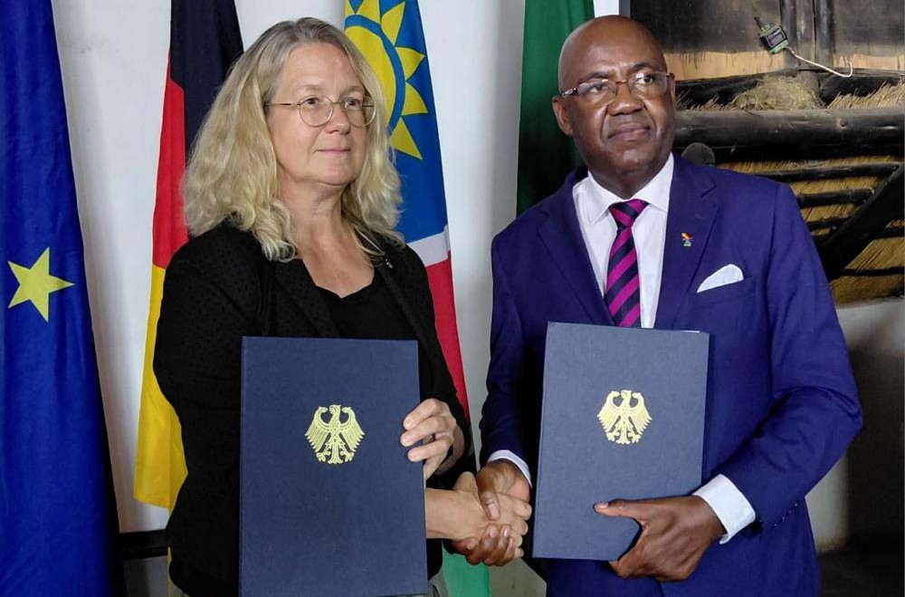 WhatsApp Image 2026-10-06 at 113001 AM - The Brief | Namibia's Leading Business & Financial News Two professionals shake hands while each holds a navy folder with a gold emblem, standing before national flags in a formal setting.