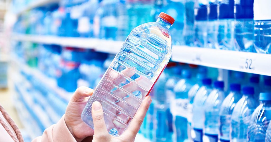 Person holding a clear plastic water bottle with a red cap in a grocery store aisle of bottled water on shelves.