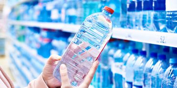 Person holding a clear plastic water bottle with a red cap in a grocery store aisle of bottled water on shelves.