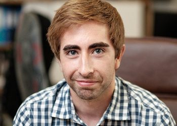 Young man with light brown hair and stubble, wearing a blue-and-white checkered shirt, looking at the camera in an office.