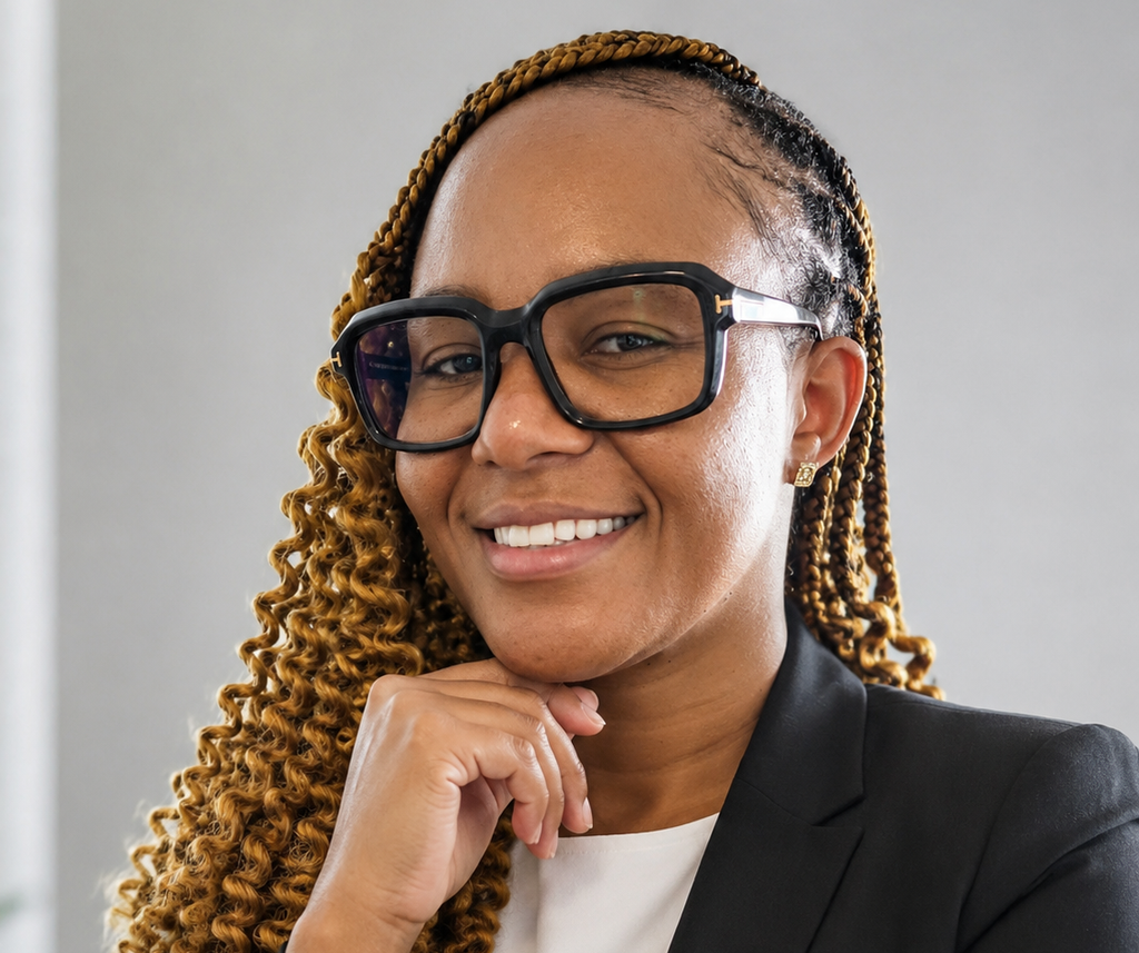 Smiling Black woman with braided hair, large black glasses, and a blazer, resting her chin on her hand.