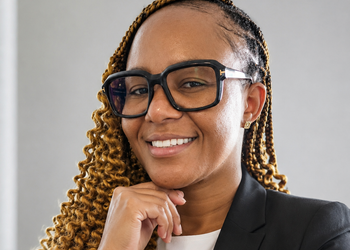 Smiling Black woman with braided hair, large black glasses, and a blazer, resting her chin on her hand.