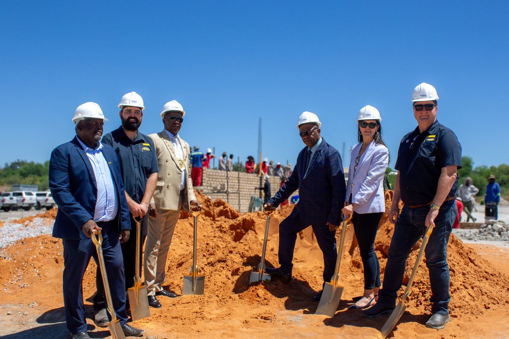 Pupkewitz Megabuild - The Brief | Namibia's Leading Business & Financial News Group of seven professionals in hard hats with shovels at a daytime groundbreaking ceremony on an orange dirt site, with a brick wall and vehicles in the background.