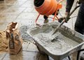 CEMENT - The Brief | Namibia's Leading Business & Financial News Construction worker mixes wet concrete in a wheelbarrow on a damp tile surface, with a cement bag nearby and a concrete mixer in the background.