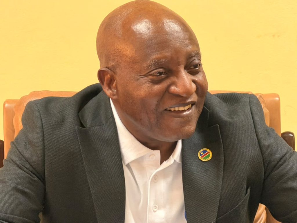 NGURARE22 - The Brief | Namibia's Leading Business & Financial News Smiling bald man in a dark blazer and white shirt, seated indoors, with a round pin on his lapel.