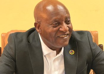 Smiling bald man in a dark blazer and white shirt, seated indoors, with a round pin on his lapel.
