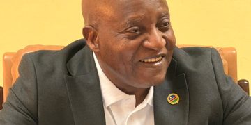 Smiling bald man in a dark blazer and white shirt, seated indoors, with a round pin on his lapel.
