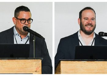 Two men in suits speak at separate wooden lecterns with laptops and microphones.