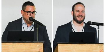 Two men in suits speak at separate wooden lecterns with laptops and microphones.