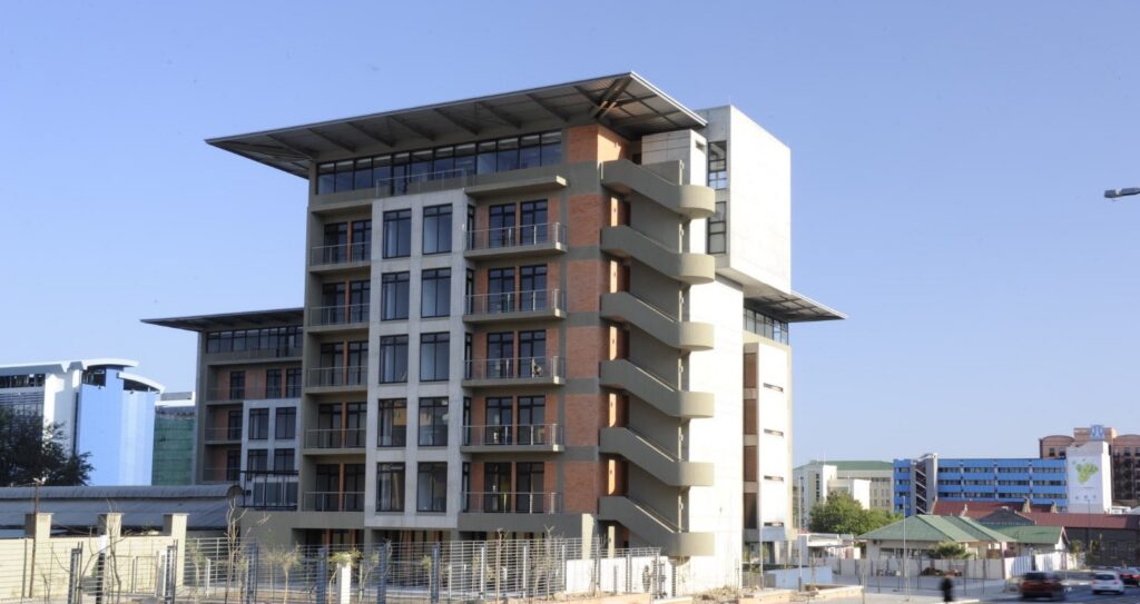 Modern six-story office building with exterior curved staircases on the right, brick and concrete façade, and large glass windows against a clear blue sky.