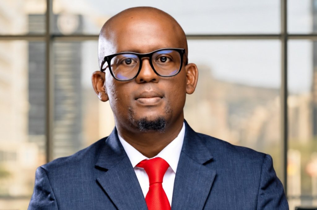 Portrait of a bald man in a navy suit and red tie, wearing black-ramed glasses, looking at the camera in an office setting